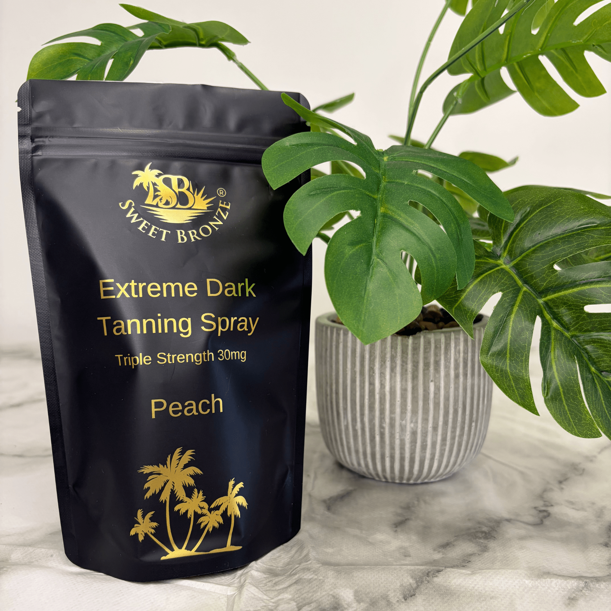 Black tanning spray package with gold text and branding, placed on a marble surface with a plant in the background.