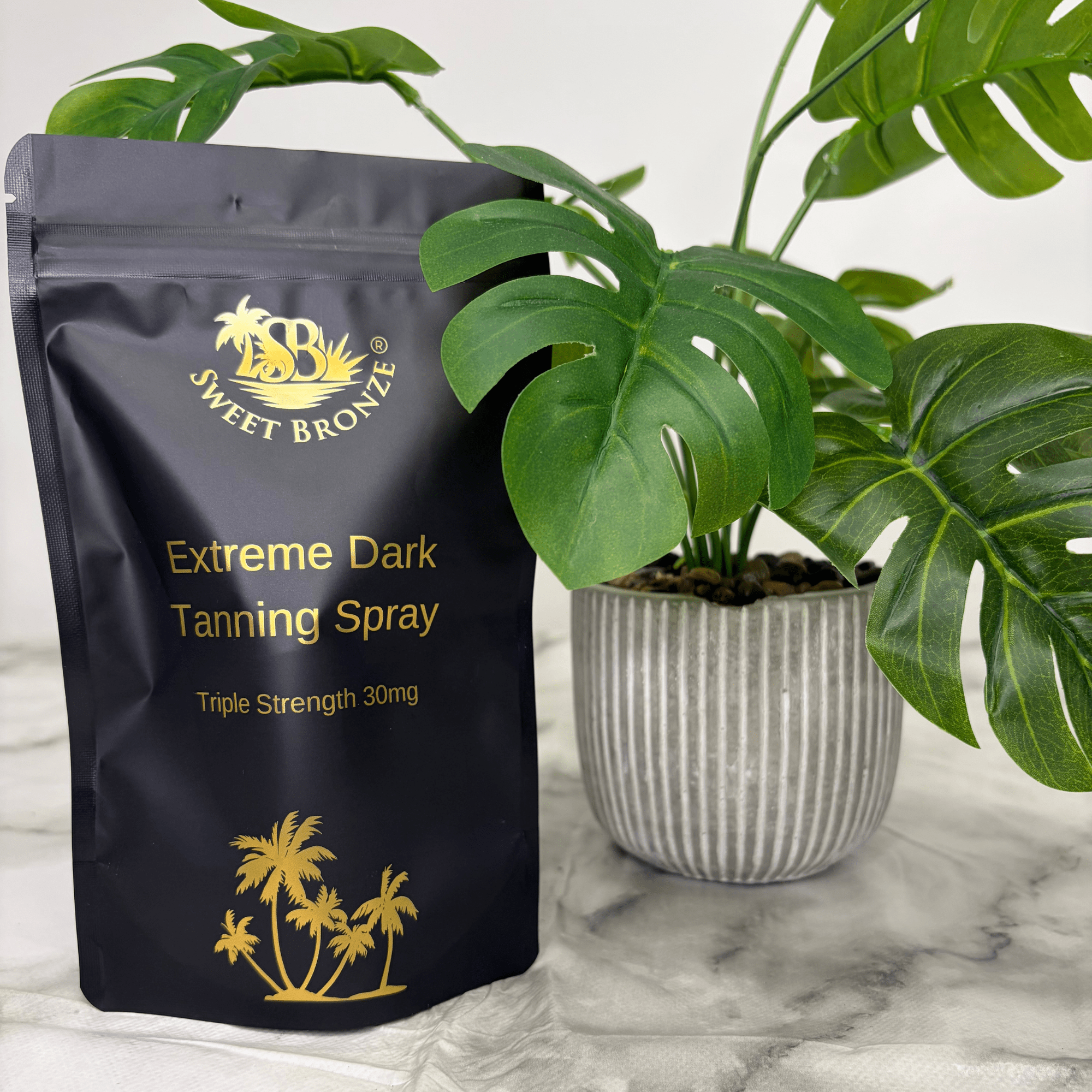 Black tanning spray package with gold text and branding next to a potted plant on a marble surface.