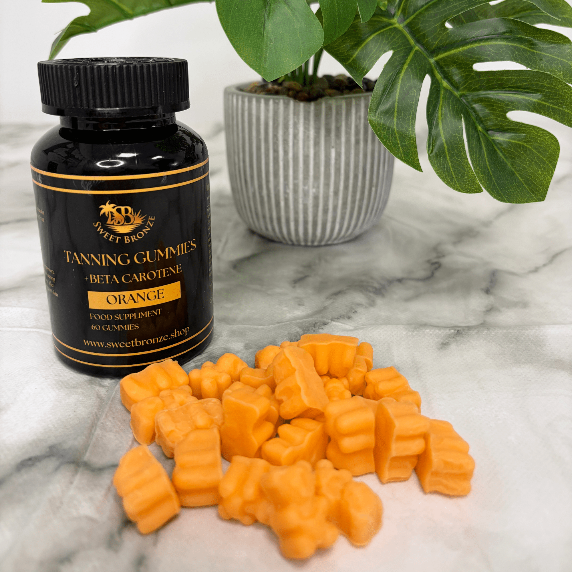 Black bottle of Sweet Bronze tanning gummies with orange gummies on a marble surface with a plant in the background.