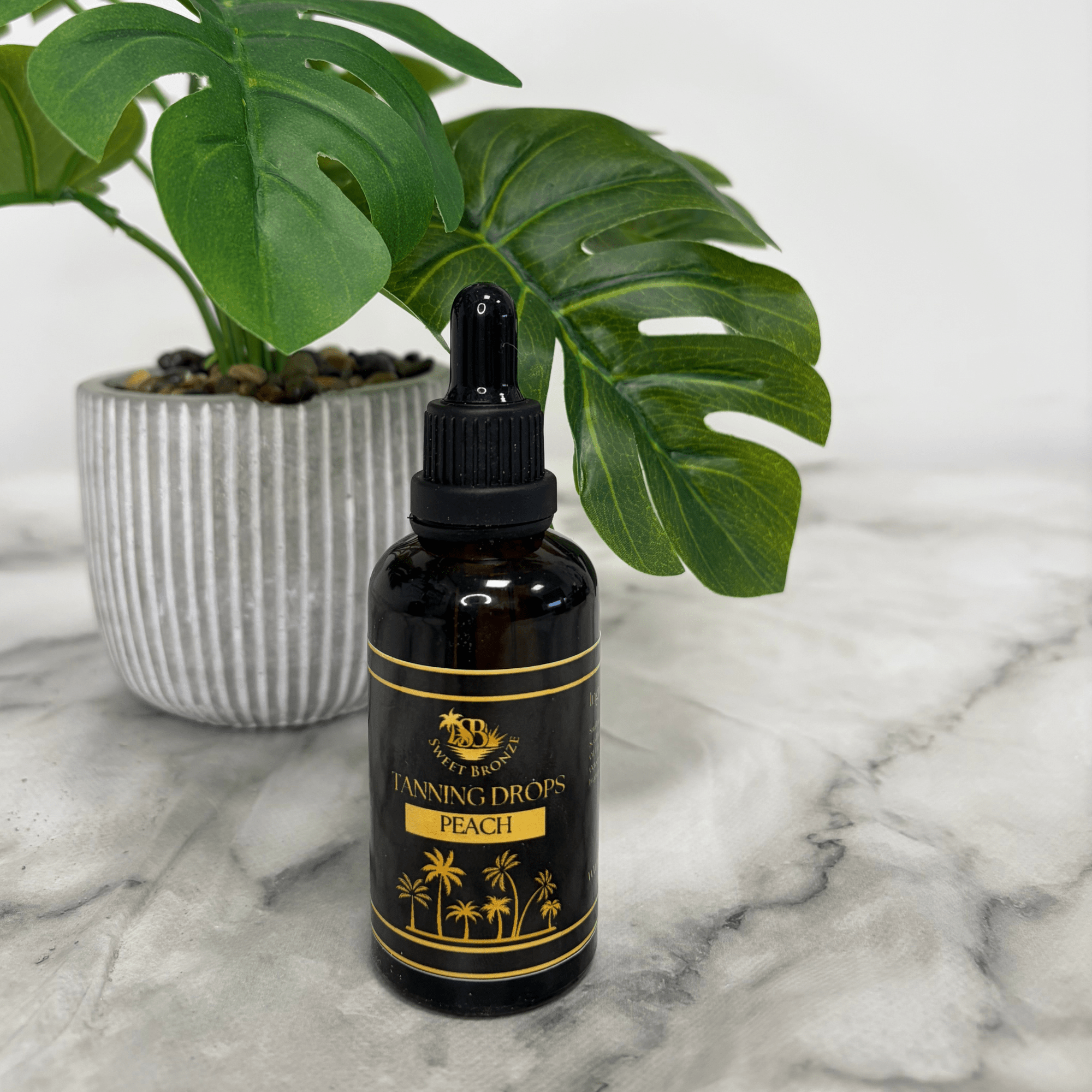 Black bottle of 'Tanning Drops Peach' on a marble surface with a plant in the background