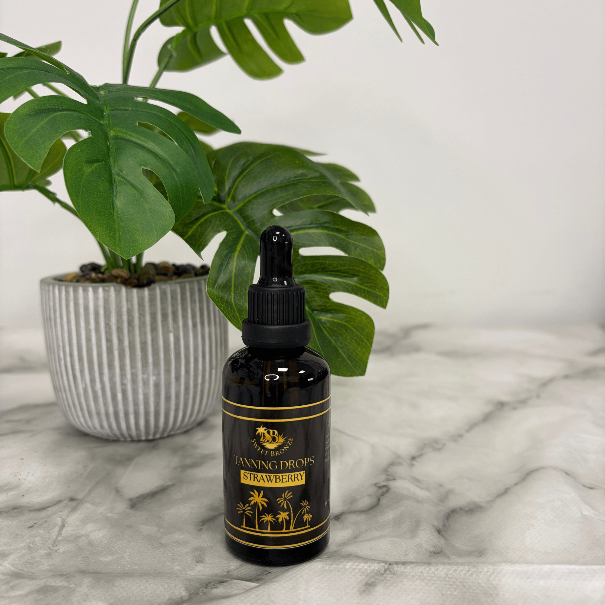 Black bottle labeled 'Tanning Drops Strawberry' on a marble surface with a plant in the background
