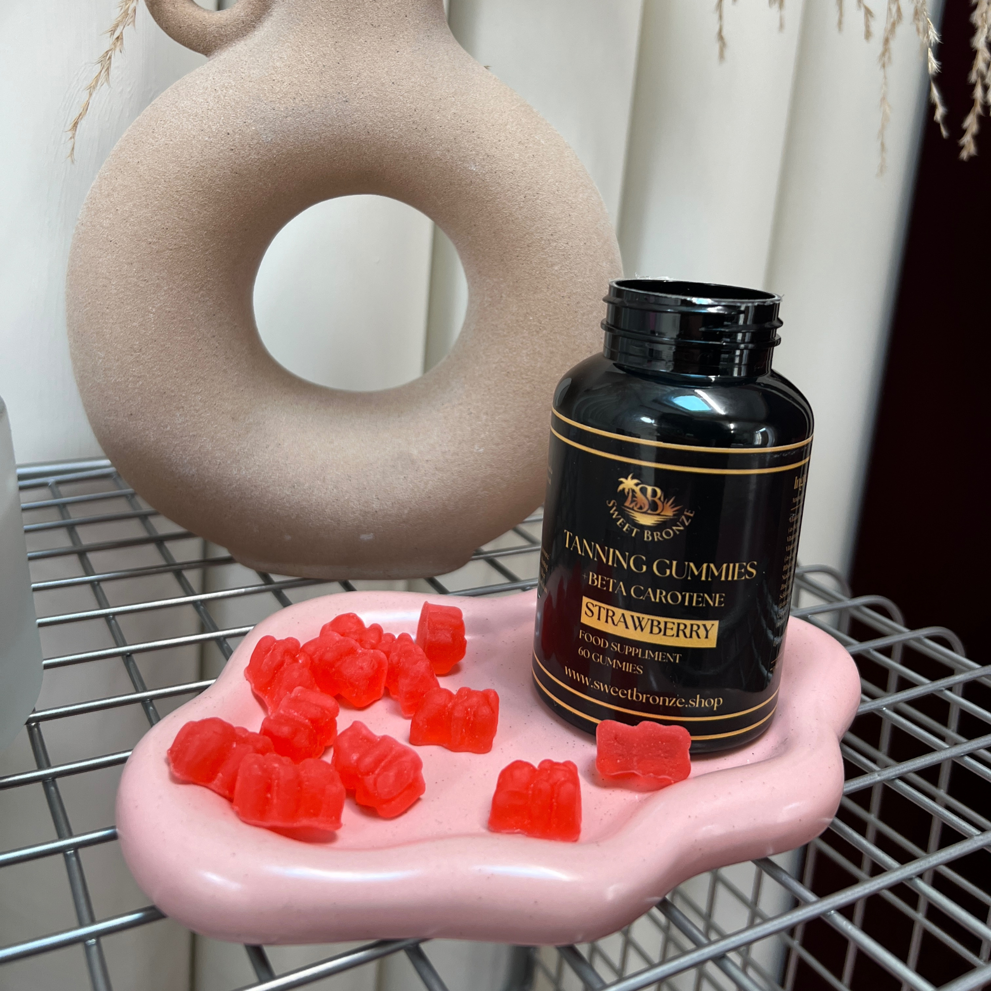 A bottle of Sweet Bronze Tanning Gummies with strawberry flavor placed on a rack, with the gummy bears scattered in front. The gummies are red, and the bottle has a black label with gold accents.