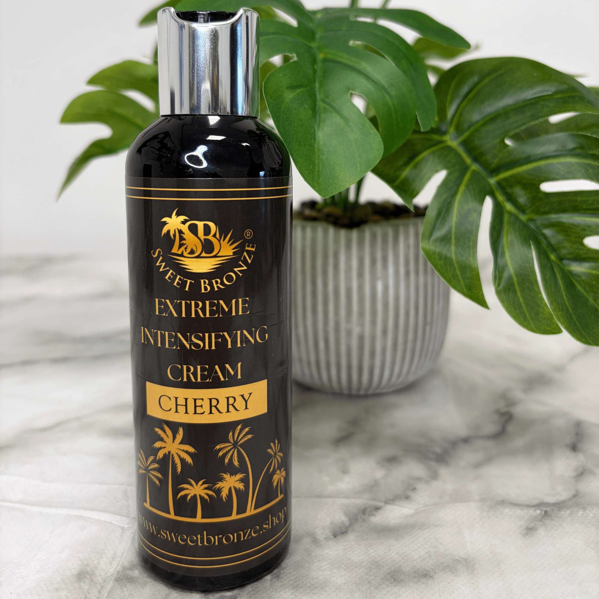 Bottle of Sweet Bronze Extreme Intensifying Cream Cherry on a marble surface with a plant in the background