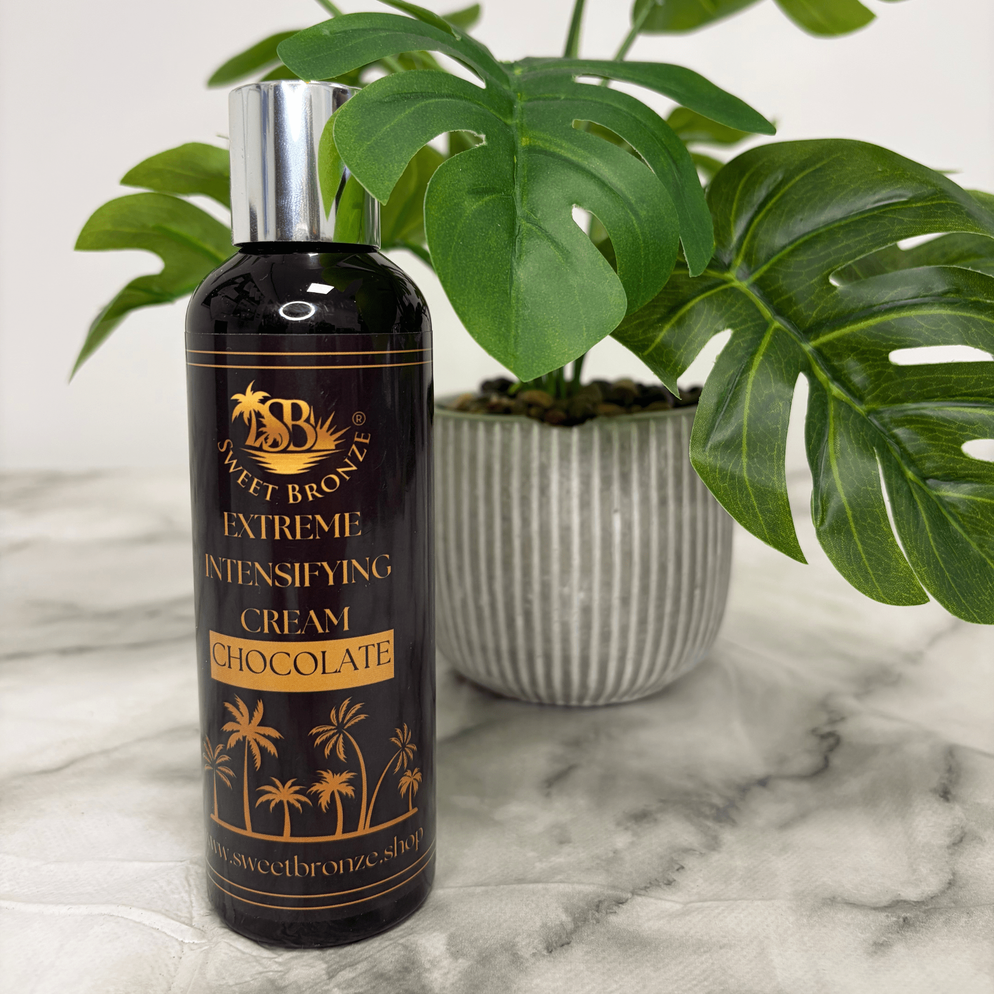 Bottle of Sweet Bronze Extreme Intensifying Cream Chocolate on a marble surface with a plant in the background