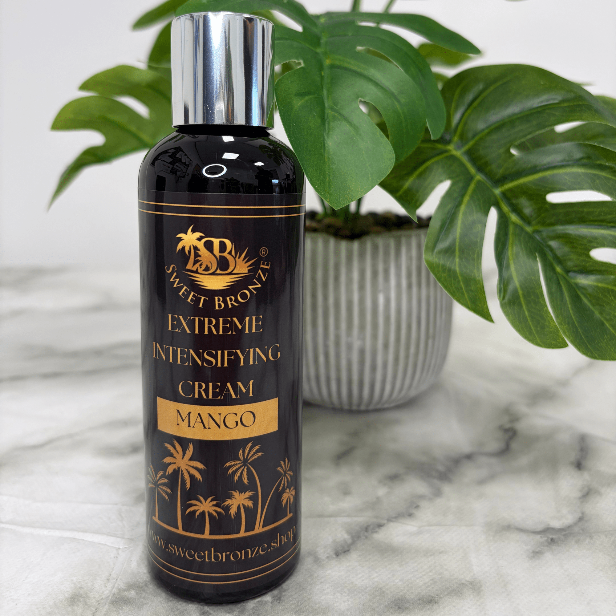 Bottle of Sweet Bronze Extreme Intensifying Cream Mango on a marble surface with a plant in the background