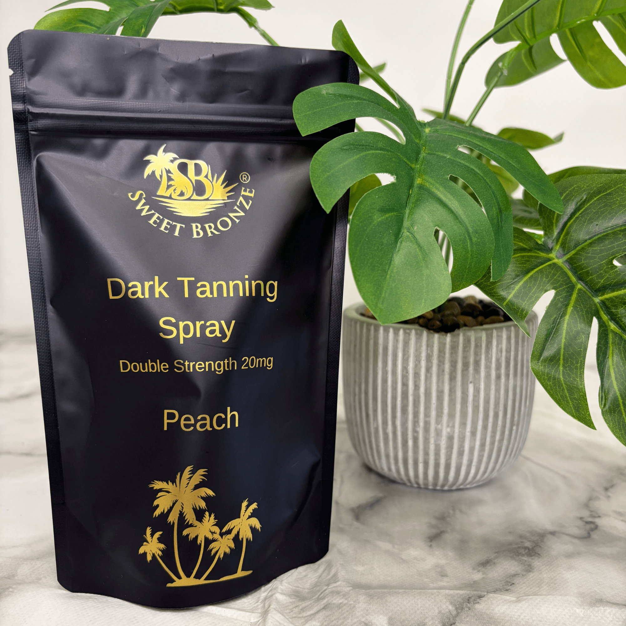 Black packaging of Sweet Bronze Dark Tanning Spray with gold text and branding, placed on a marble surface with a plant in the background.