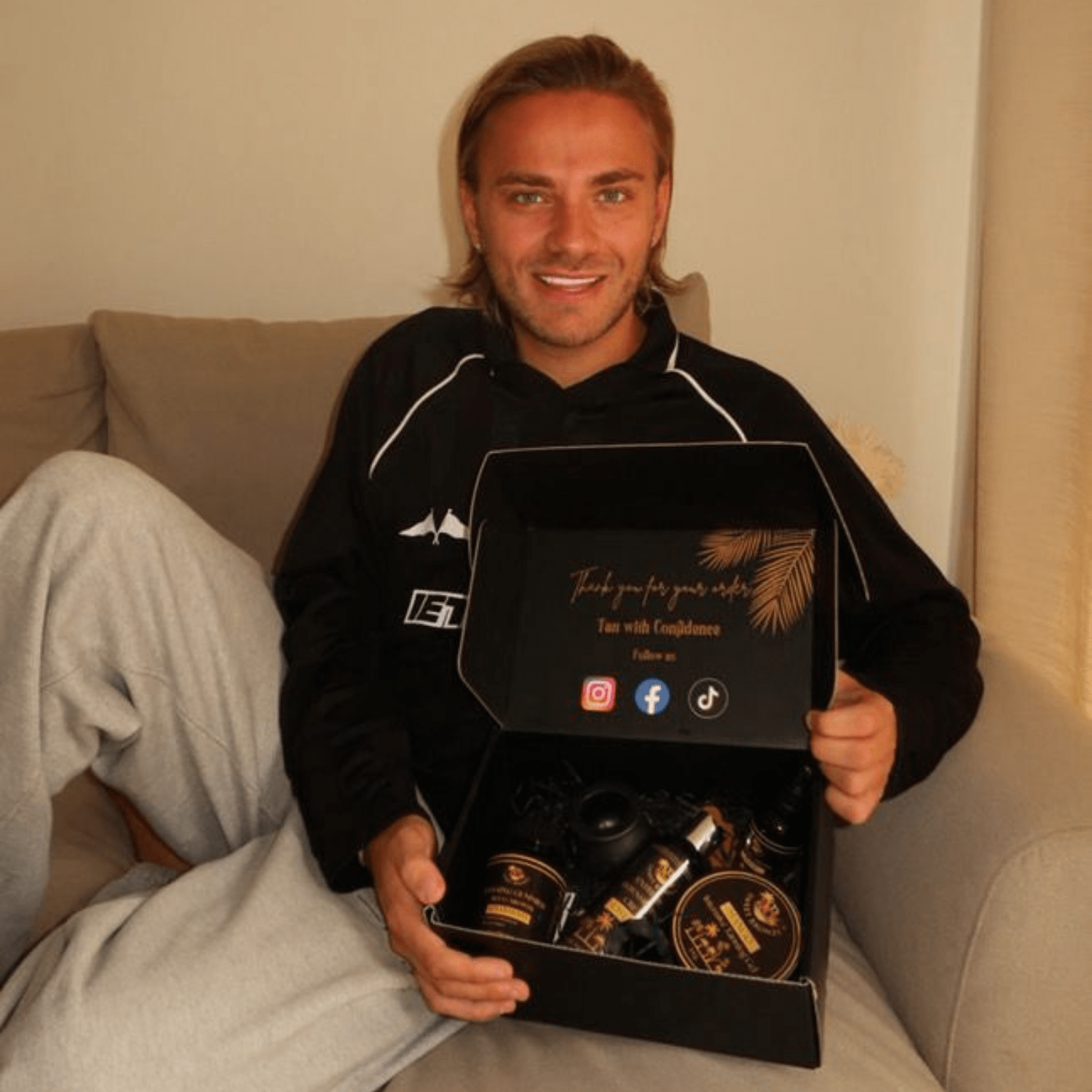 Man, Freddie Bentley, holding a black gift box with tanning products on a couch
