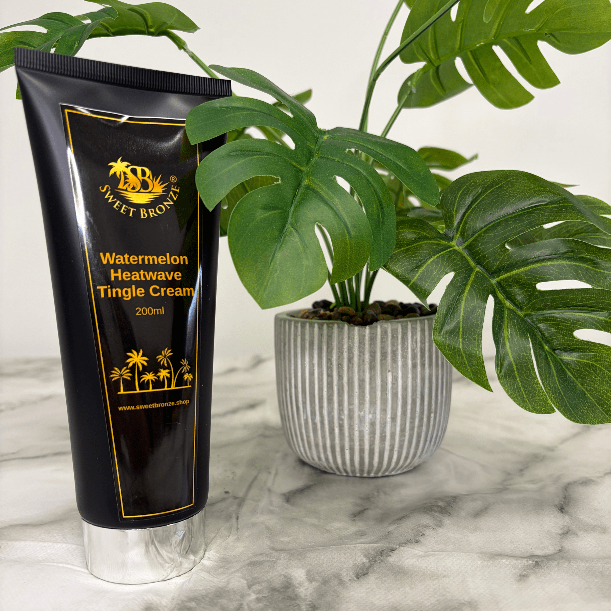 Black tube of Sweet Bronze Watermelon Heatwave Tingle Cream on a marble surface with a plant in the background